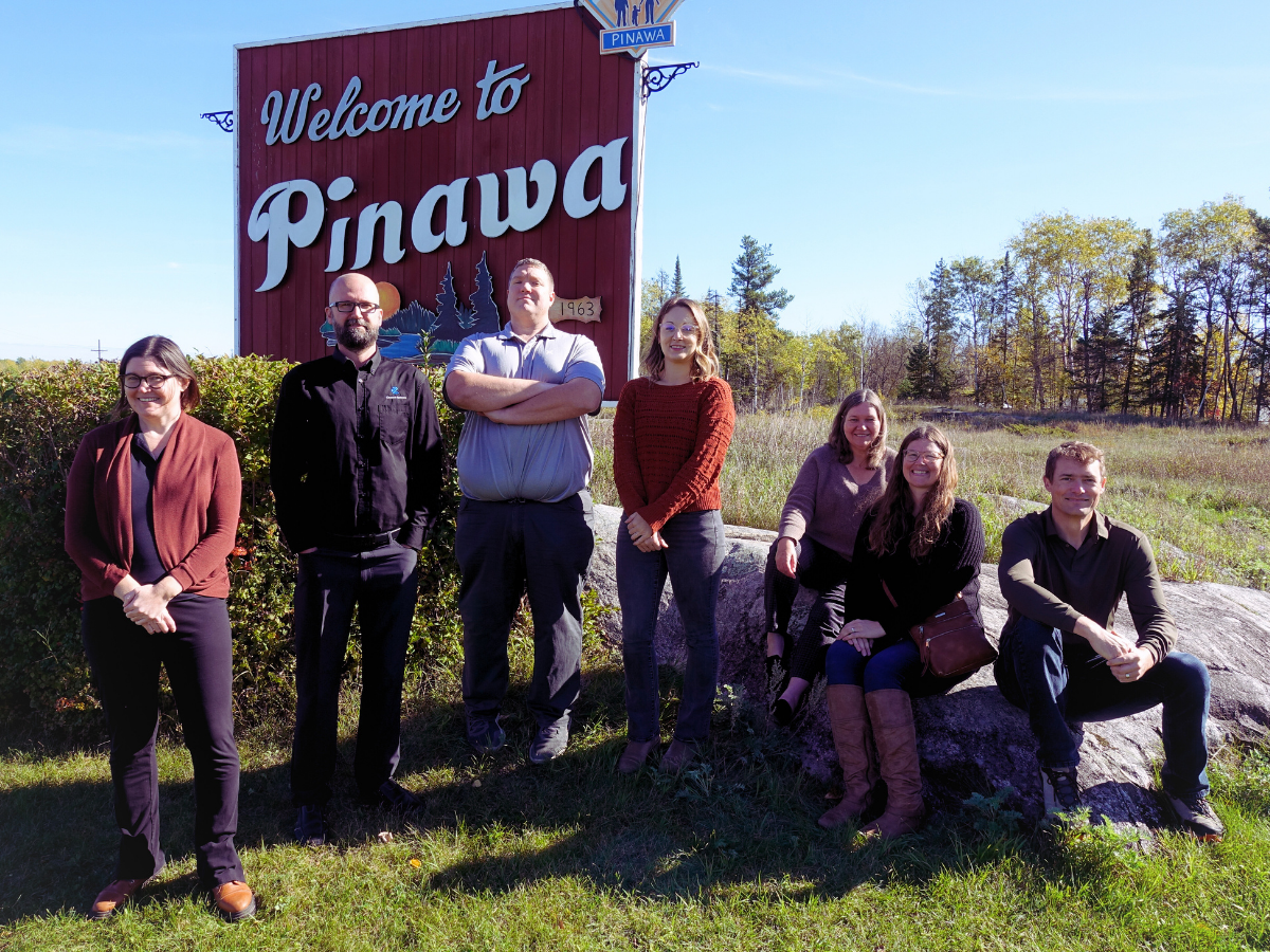 Channel Systems team posing outdoors in the summer in front of a Welcome to Pinawa sign.